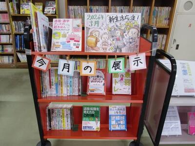 本を載せたカートの写真。「今月の展示」「新生活応援！」という掲示がなされています。
